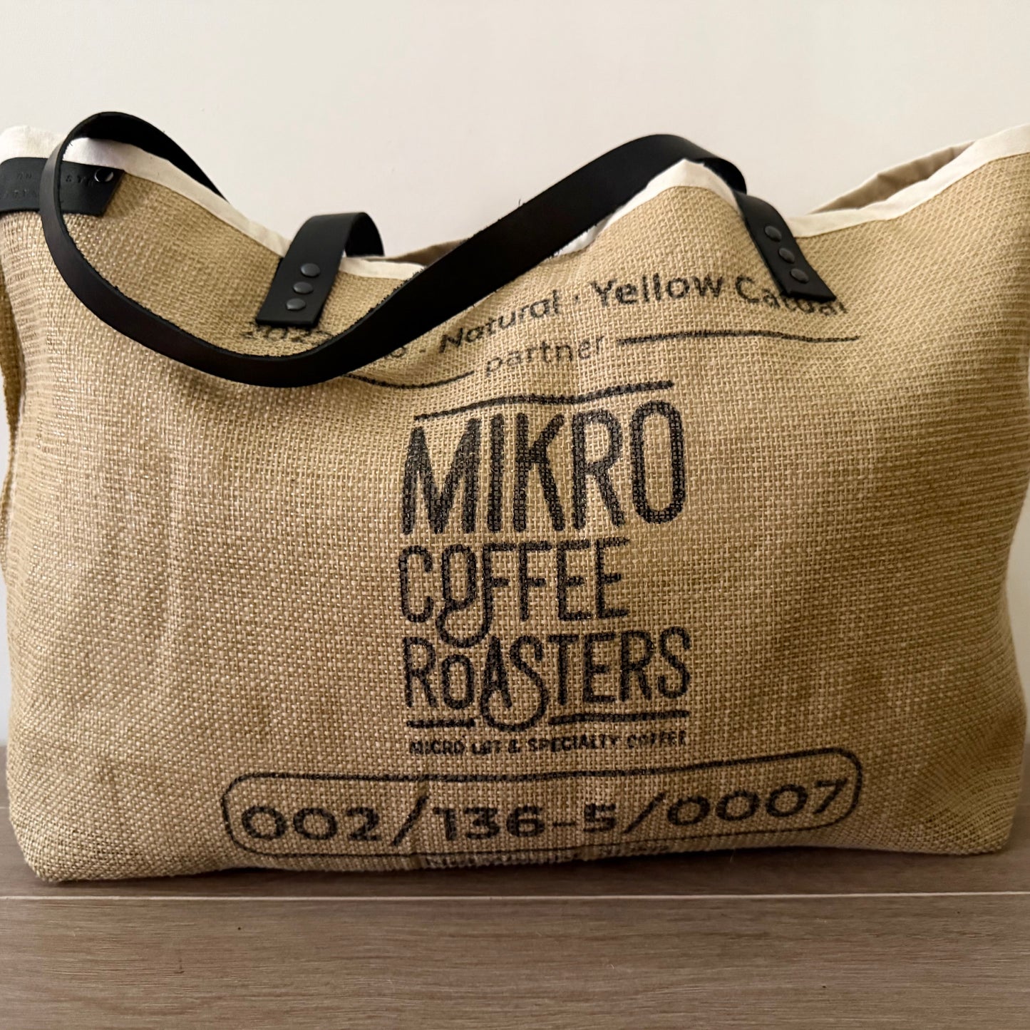 A beige upcycled tote bag with black leather handles and black text that reads 'MIKRO COFFEE ROASTERS' along with a numerical code, indicating its unique design.